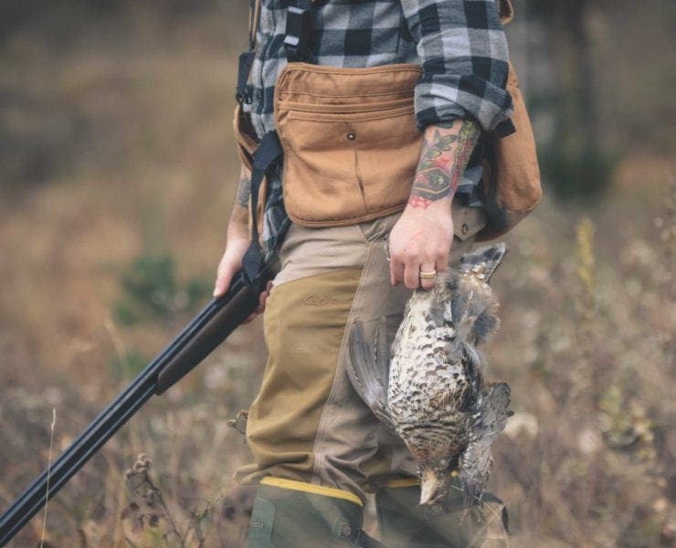 A hunter carries a grouse in the woods with a shotgun.