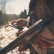 A hunter looks at a smart phone while holding a shotgun