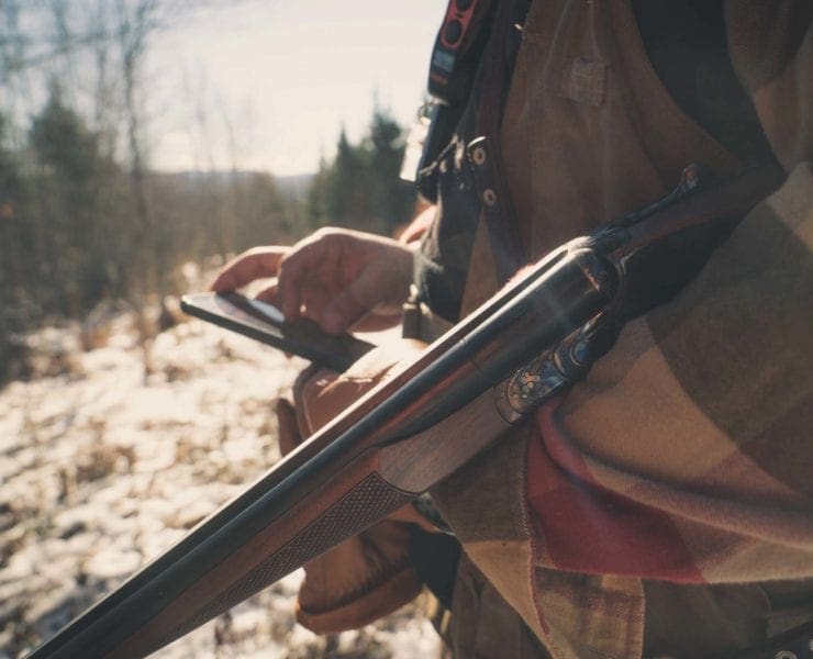 A hunter looks at a smart phone while holding a shotgun