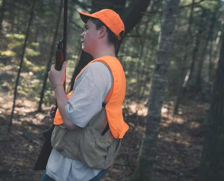 A young hunter holds a youth shotgun wearing blaze orange in the woods