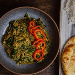 Palak Pheasant rests in a bowl next to a side of naan bread.