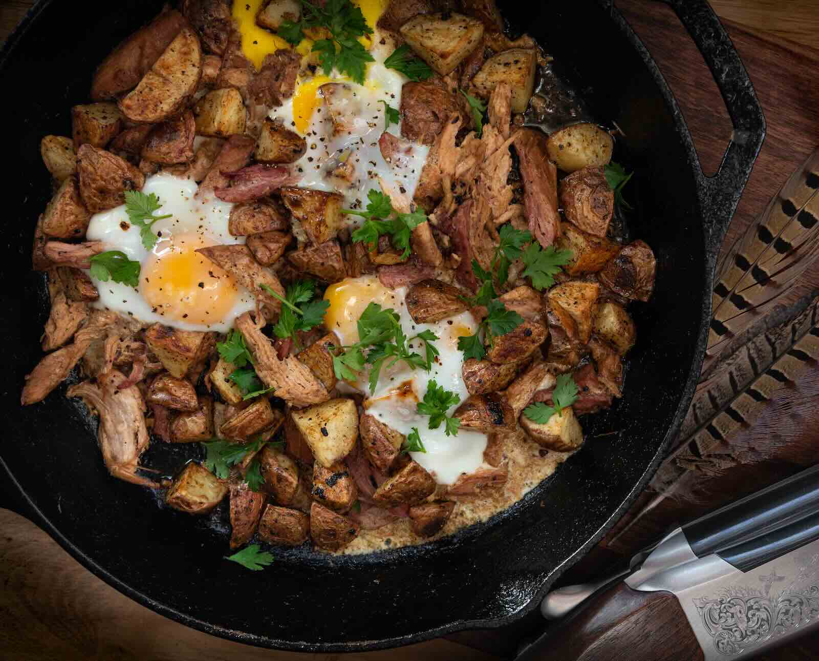 Corned pheasant hash with fried egg and red potatoes in a cast iron skillet with an accent of pheasant feathers