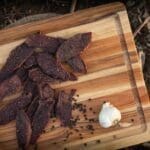Dehydrated goose jerky on a wood cutting board with a head of garlic and black peppercorns