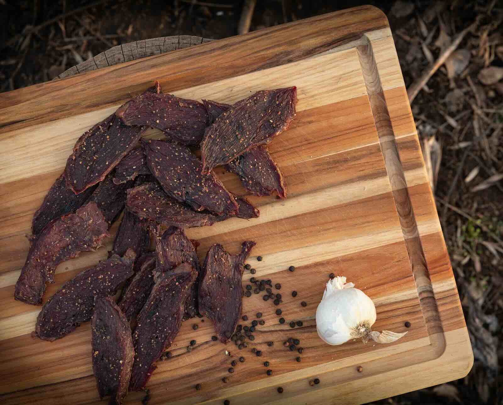 Dehydrated goose jerky on a wood cutting board with a head of garlic and black peppercorns