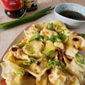 Quail-filled wonton dumplings topped with chili crisp and green onions on a white plate with soy sauce
