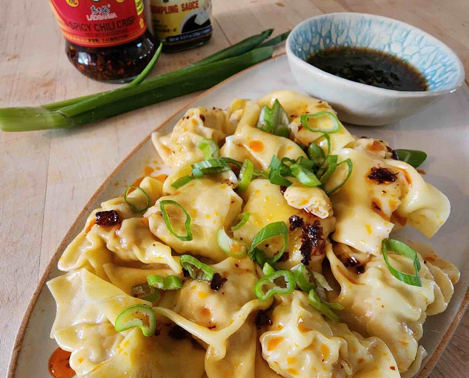 Quail-filled wonton dumplings topped with chili crisp and green onions on a white plate with soy sauce