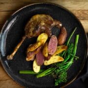 One leg of confit goose served on a black plate with potatoes and steamed broccoli, on a wood plank background