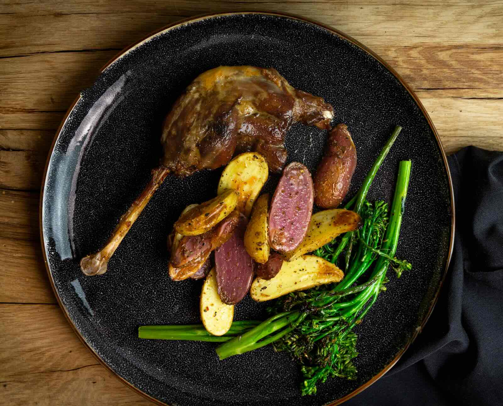 One leg of confit goose served on a black plate with potatoes and steamed broccoli, on a wood plank background