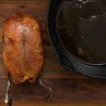 A whole roasted duck with crispy skin sits on a wooden table next to a cast iron pan