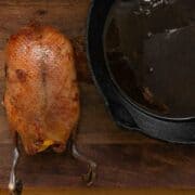 A whole roasted duck with crispy skin sits on a wooden table next to a cast iron pan