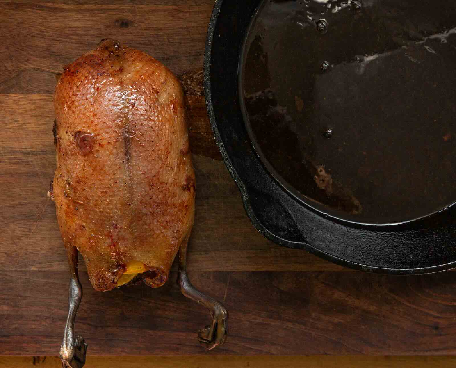 A whole roasted duck with crispy skin sits on a wooden table next to a cast iron pan