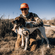 A man holding a bird hugs a dog in the desert.