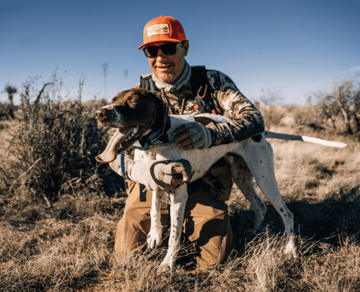 A man holding a bird hugs a dog in the desert.