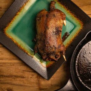 A browned, roasted whole mallard duck is served on a green plate next to the cast iron skillet it was cooked in.