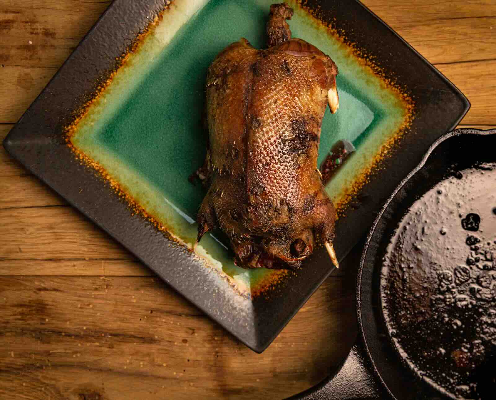 A browned, roasted whole mallard duck is served on a green plate next to the cast iron skillet it was cooked in.