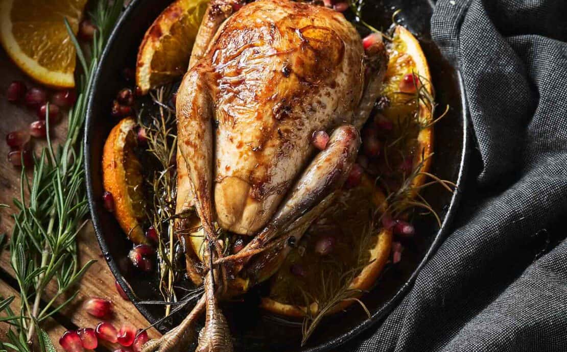 A whole, roasted ruffed grouse is served in a cast iron pan with a background of herbs, fruit, and linen