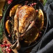 A whole, roasted ruffed grouse is served in a cast iron pan with a background of herbs, fruit, and linen