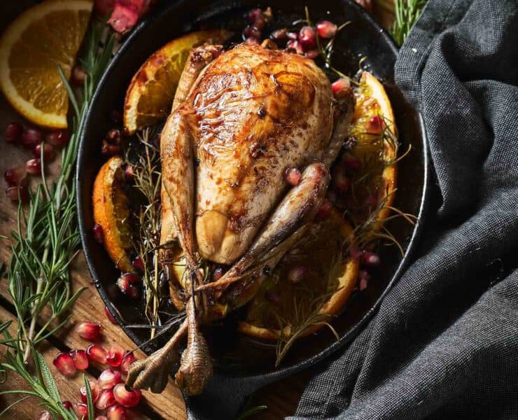 A whole, roasted ruffed grouse is served in a cast iron pan with a background of herbs, fruit, and linen