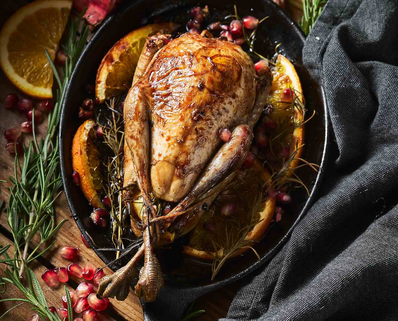 A whole, roasted ruffed grouse is served in a cast iron pan with a background of herbs, fruit, and linen