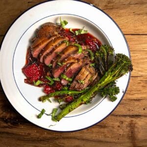 Sliced duck breast served with a raspberry sauce on a white plate with a side of steamed broccoli