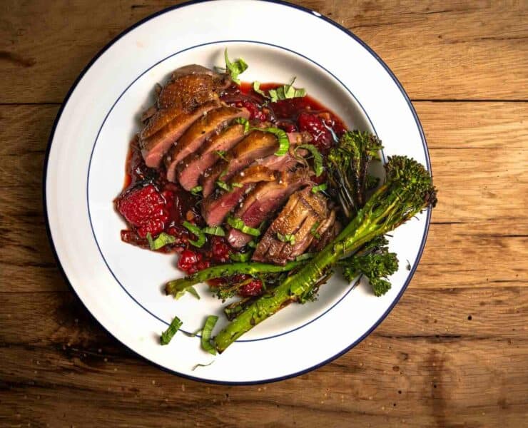 Sliced duck breast served with a raspberry sauce on a white plate with a side of steamed broccoli