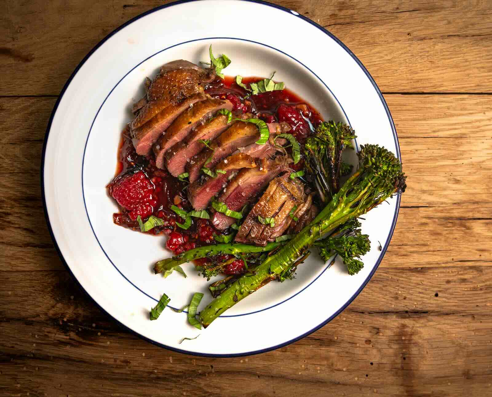 Sliced duck breast served with a raspberry sauce on a white plate with a side of steamed broccoli