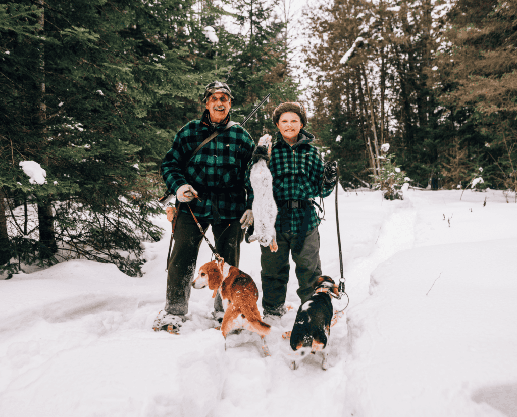 An old man and a young man stand in the snow with their two beagles and a dead snowshoe hare.