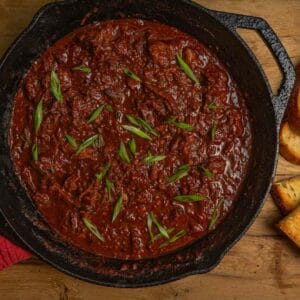 A cast iron skillet filled with goose leg chili sits on a wooden surface with slices of toasted garlic bread