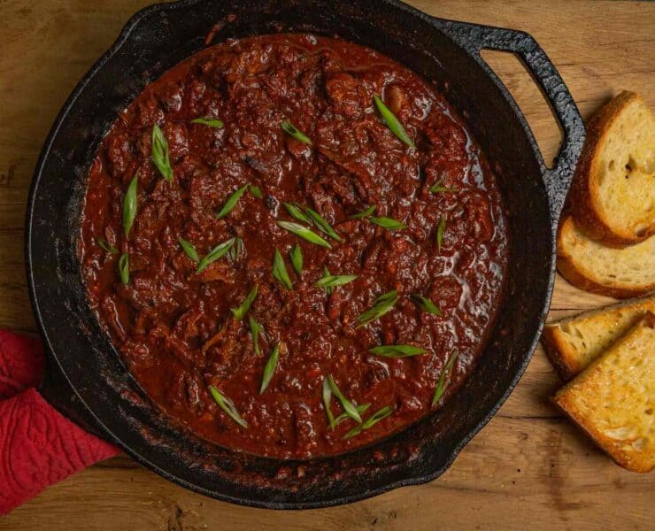 A cast iron skillet filled with goose leg chili sits on a wooden surface with slices of toasted garlic bread