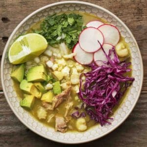 A white bowl of quail pozole topped with avocado, red cabbage, radish, cilantro, and lime wedges.