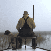 A man sits on the edge of a boat with a gun and his dog.