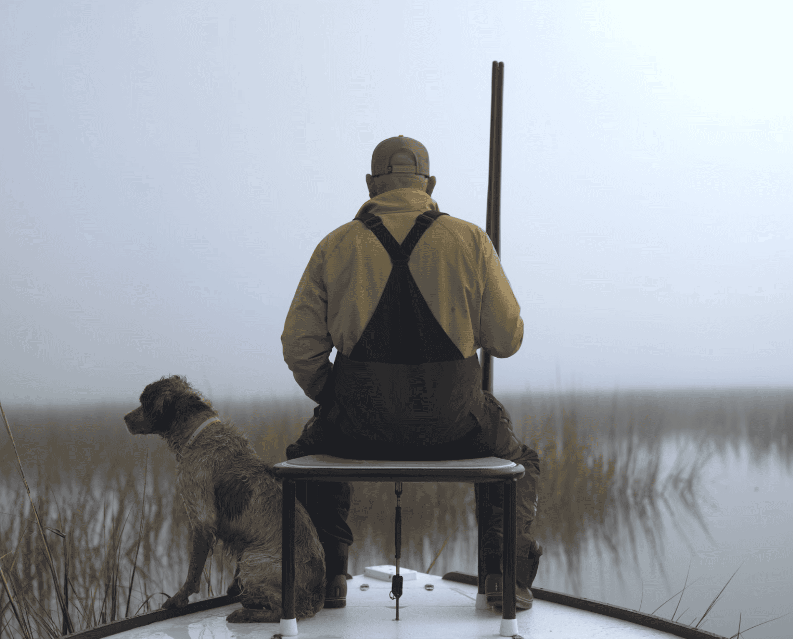 A man sits on the edge of a boat with a gun and his dog.