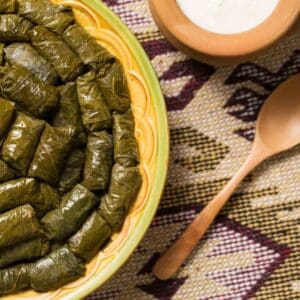 A plate of rolled dolmas served on a woven placemat with a wooden spoon and a bowl of white sauce