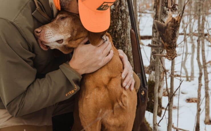 A Vizsla with one front leg hugs his owner in the woods next to a hanging dead American woodcock.