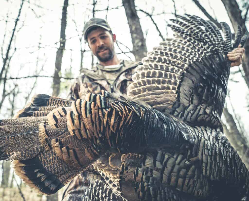 A man wearing camouflage and holding a dead male wild turkey.
