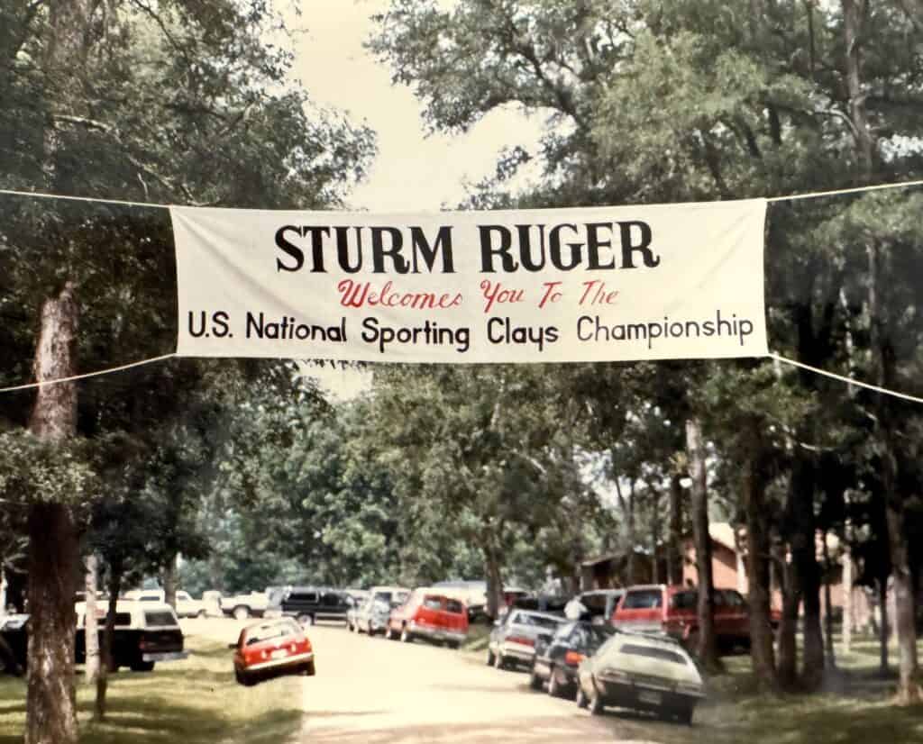 A banner that states "Sturm Ruger welcomes you to the U.S. National Sporting Clays Championship" hunt over a road filled with parked cars in 1985.
