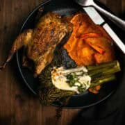 A whole roasted chukar, sweet potato puree, and broccoli are served on a plate with a fork and knife on a dark wooden background