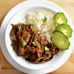 Braised and shredded turkey meat is coated in a caramel sauce and served with white rice and cucumber slices in a white bowl