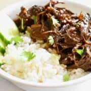 Braised and shredded turkey meat is coated in a caramel sauce and served with white rice and cucumber slices in a white bowl