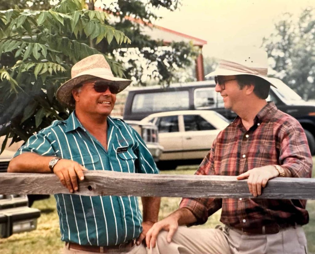 Jay Herbert and Bob Davis standing along a fencepost with cars in the background.