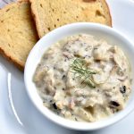 A white bowl filled with chunky cream of mushroom soup served with two slices of bread on a white plate