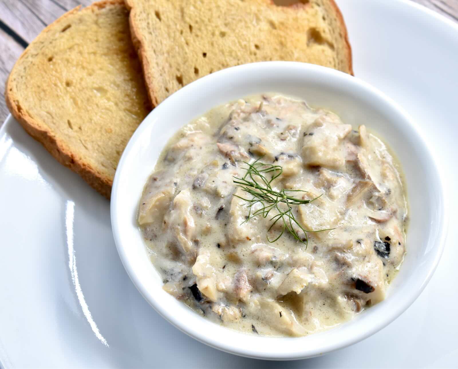 A white bowl filled with chunky cream of mushroom soup served with two slices of bread on a white plate