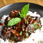 Roasted muskrat meat coated in a brown sauce, served atop white rice in a black bowl and garnished with Thai basil