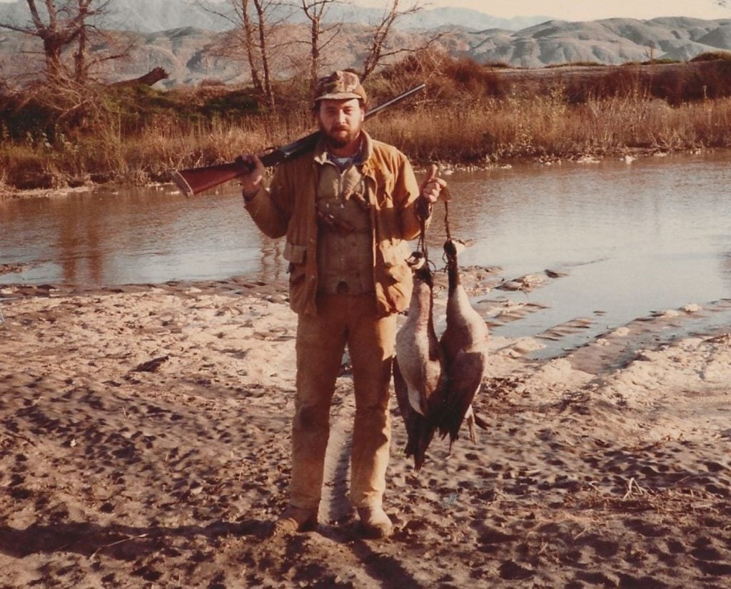 The author William Murray holding two geese and a shotgun along a river.
