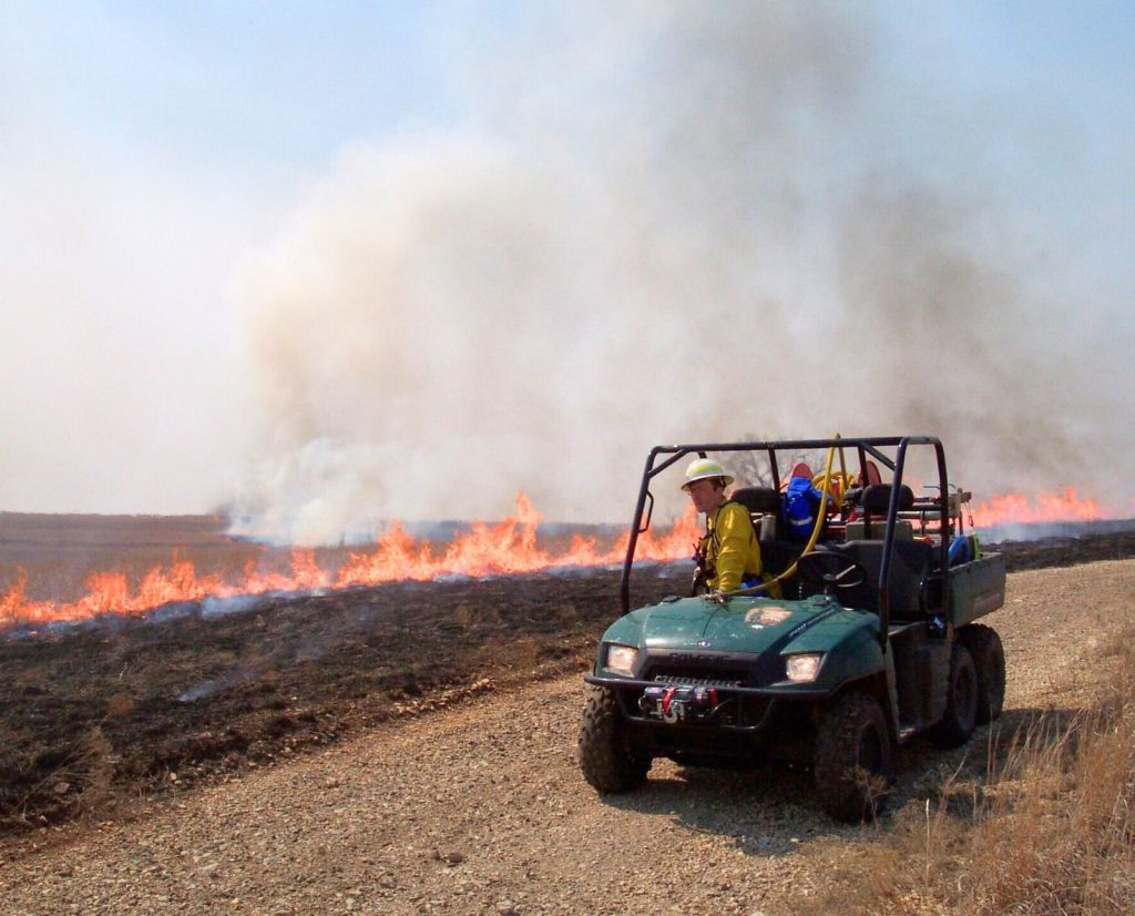 A firefighter looks at a controlled burn from a side-by-side with a water tank and hoses.