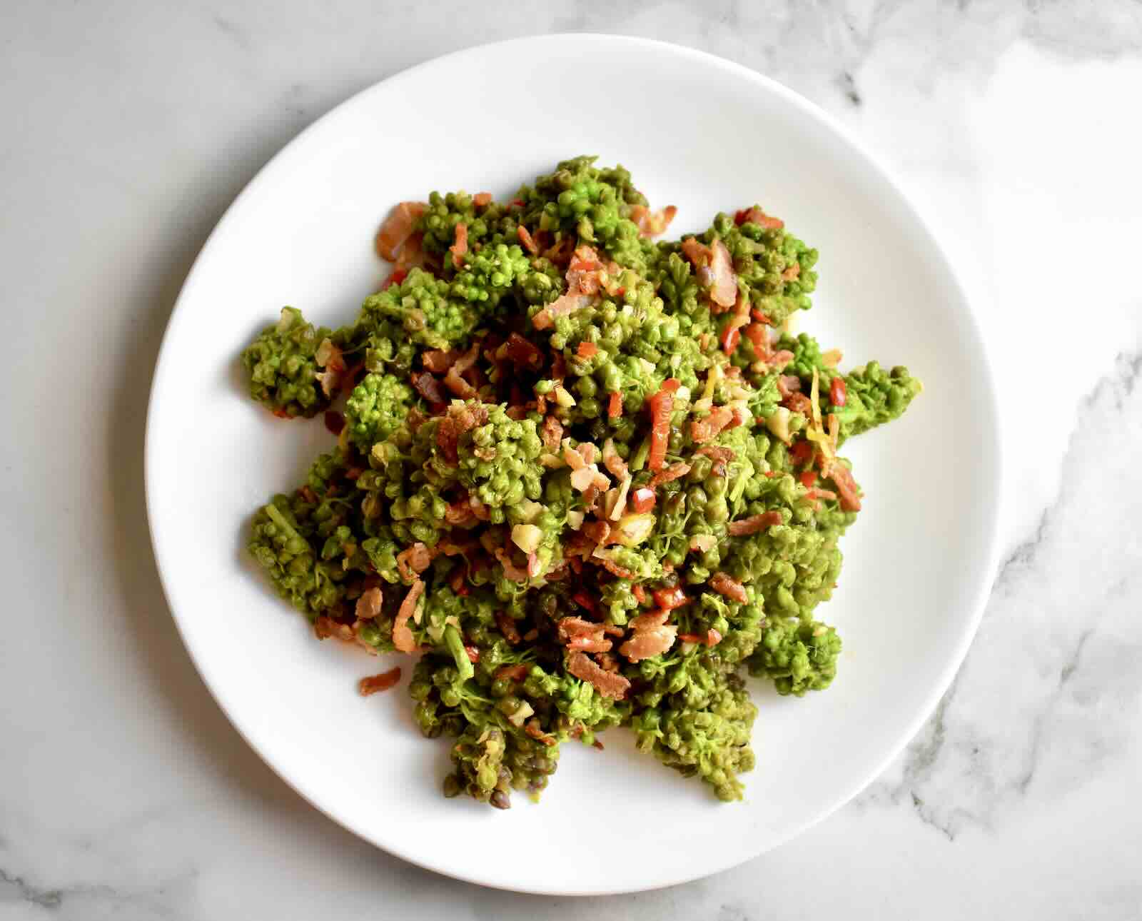 Cooked milkweed buds with bacon, garlic, chilies, and anchovies served on a white plate atop a marble countertop