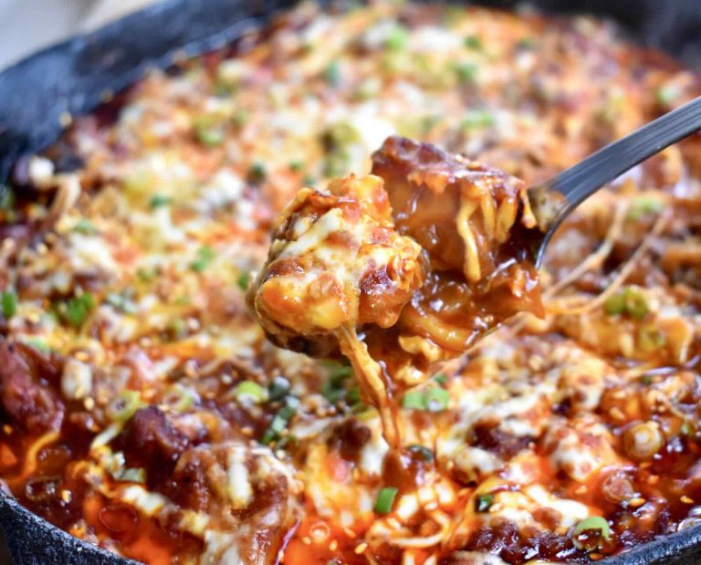 A serving of spicy, cheesy rabbit meat cooked with Korean spices is held in a spoon above a cast iron skillet