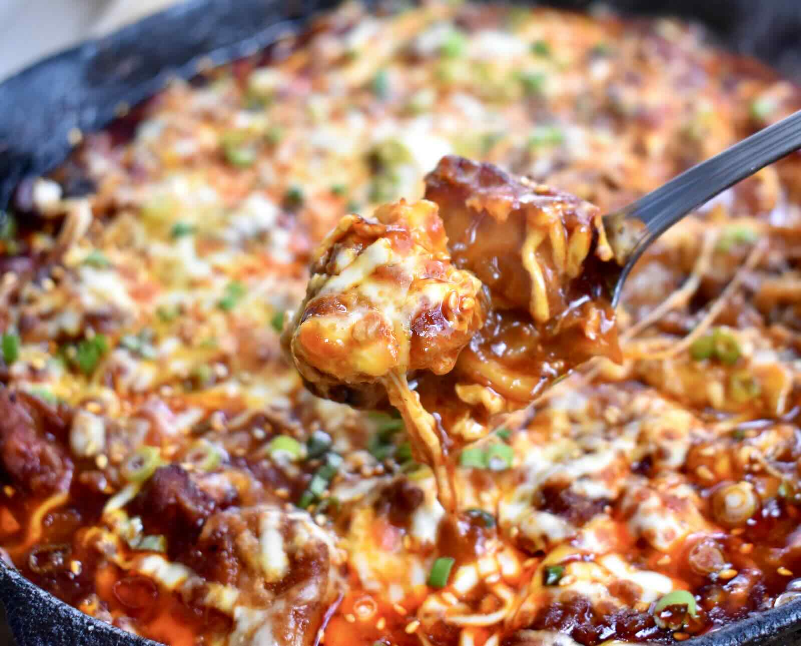 A serving of spicy, cheesy rabbit meat cooked with Korean spices is held in a spoon above a cast iron skillet